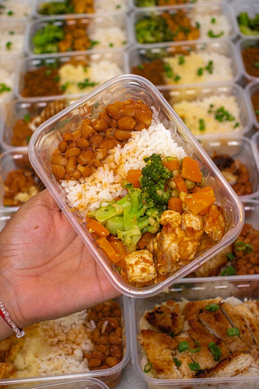 Mão segurando uma marmita de arroz, feijão, frango e legumes, com dezenas de marmitas prontas ao fundo
